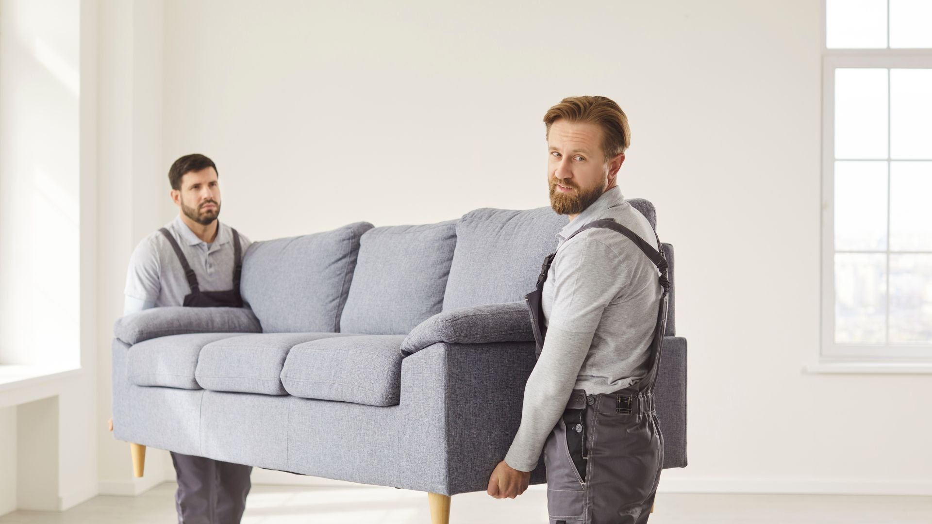 Two movers carefully carrying a gray couch in a bright room