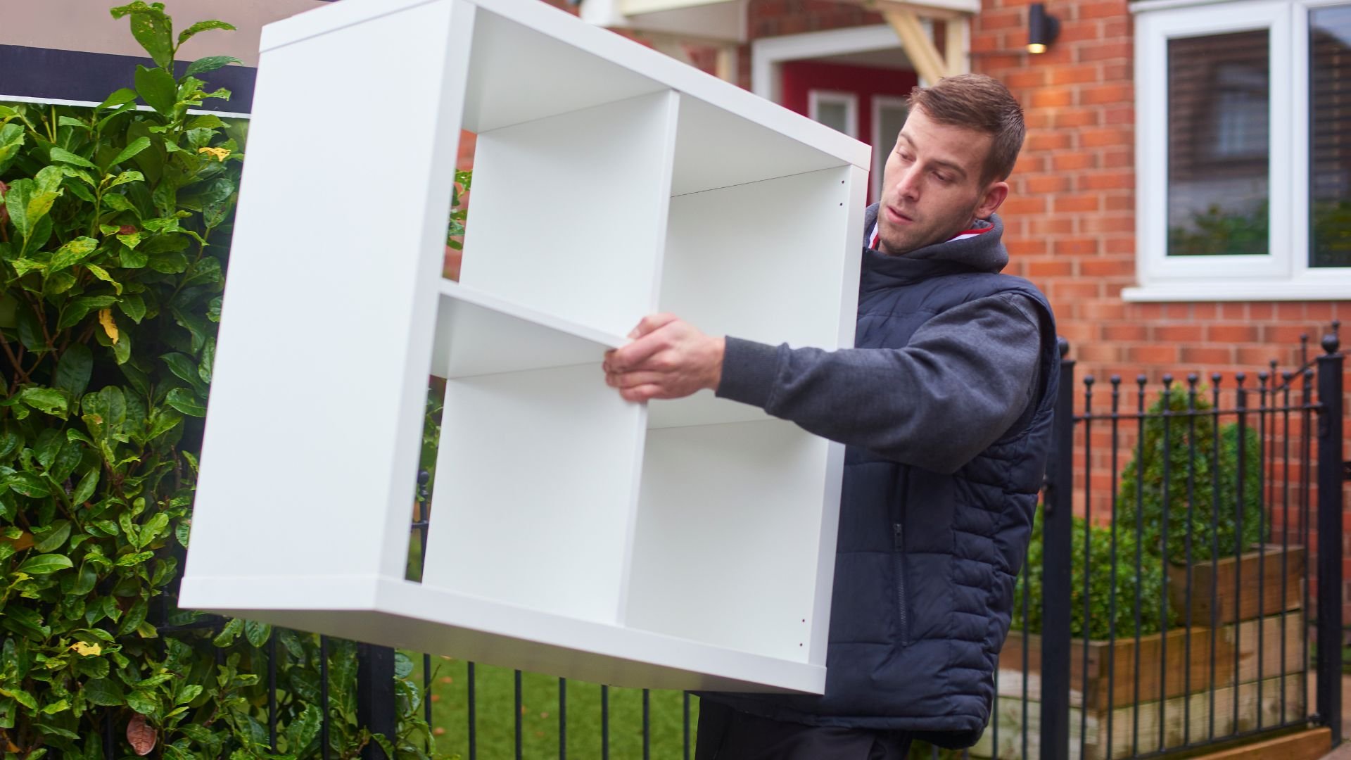 Person carrying white shelving unit outdoors near brick house and garden