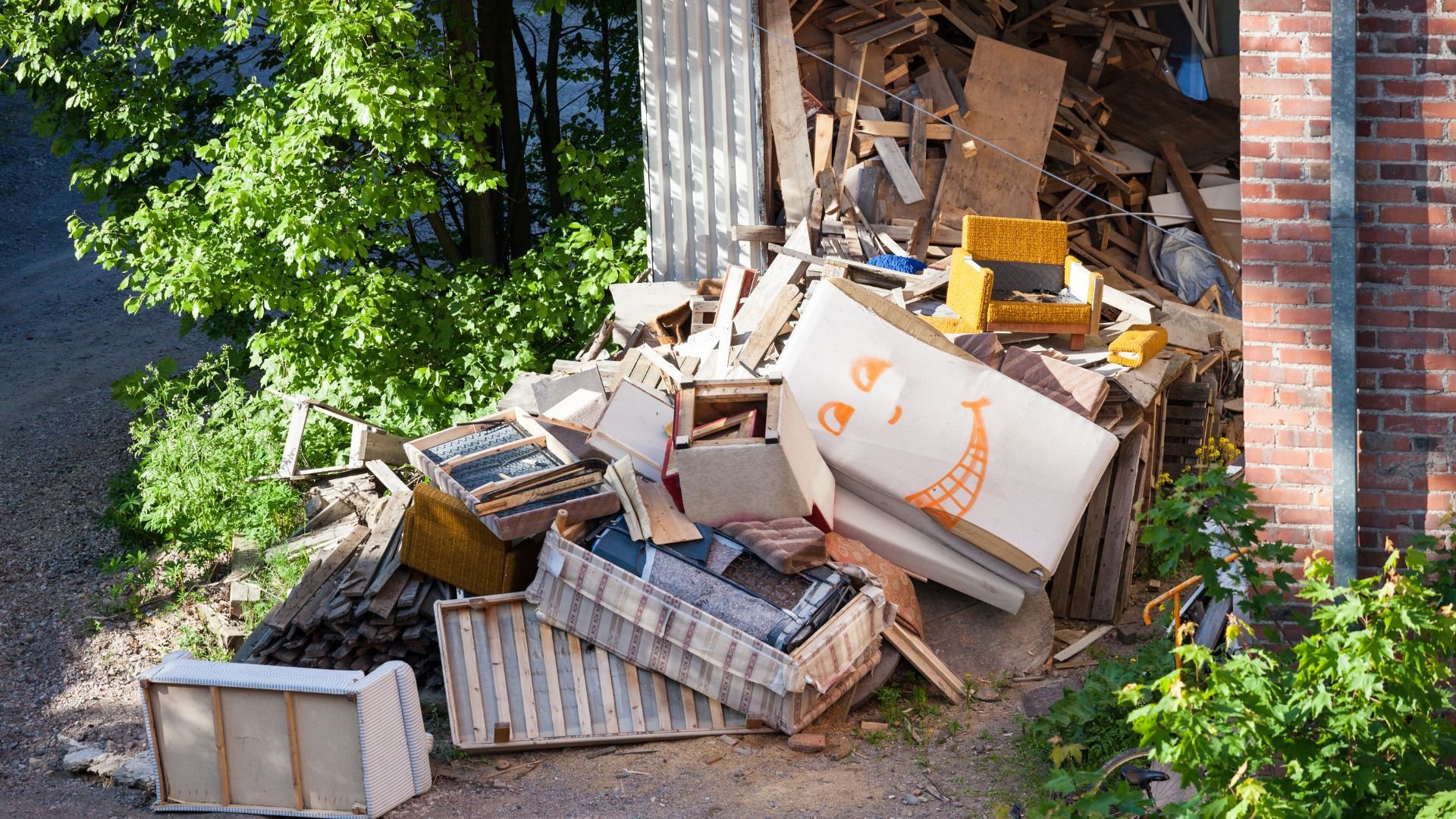 Cluttered pile of wood, furniture, and debris near brick wall and trees